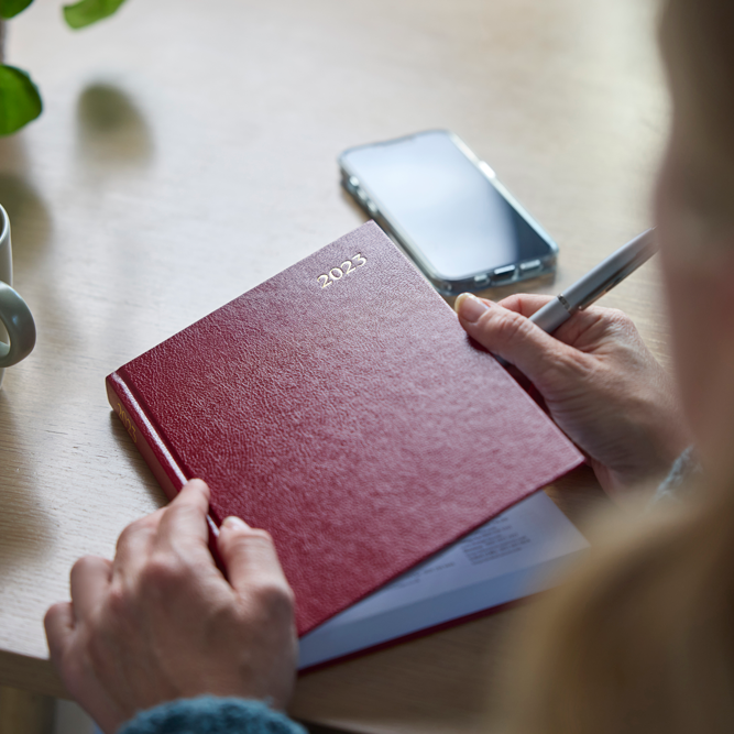 Person holding a pen about to open 2023 diary. Mug of coffee and mobile phones also on desk.